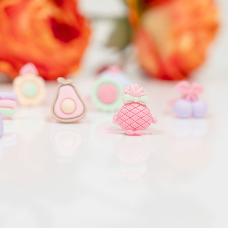 May include: A set of colorful fruit-shaped hair clips, including a pineapple, avocado, and a pair of cherries. The clips are made of plastic and have a glossy finish.
