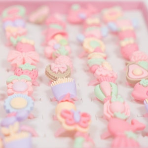 May include: A collection of colorful plastic rings with various designs, including flowers, butterflies, hearts, and strawberries. The rings are arranged in rows on a pink background.