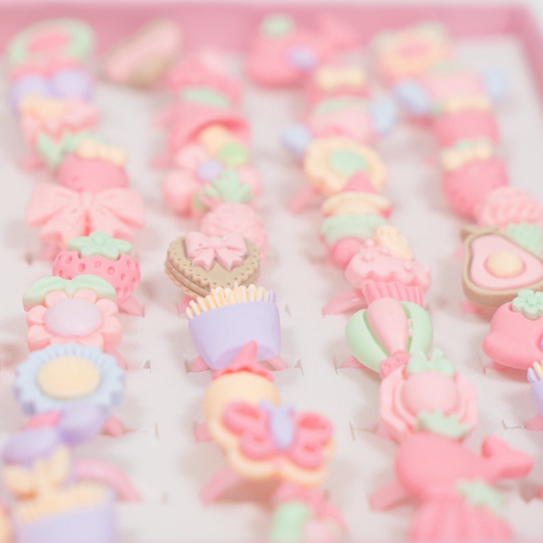 May include: A collection of colorful plastic rings with various designs, including flowers, butterflies, hearts, and strawberries. The rings are arranged in rows on a pink background.