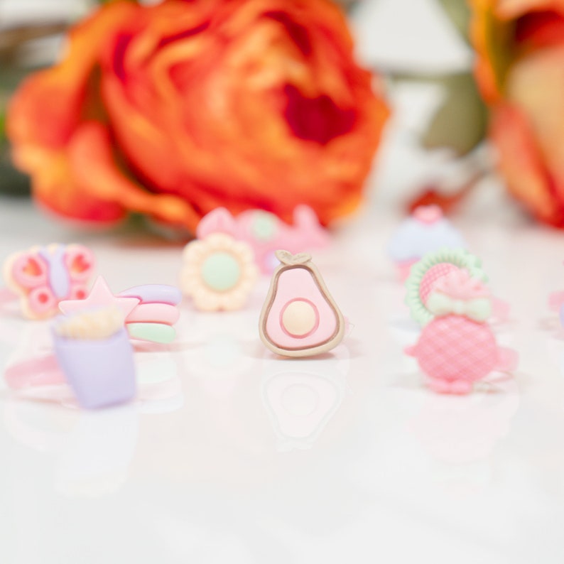 May include: A collection of colorful, whimsical rings featuring various designs, including a butterfly, a star, a rainbow, a flower, a cupcake, a pineapple, and an avocado. The rings are made of plastic and are perfect for adding a touch of fun to any outfit.