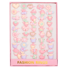 May include: A pink box with a white interior containing 36 colorful plastic rings with various designs, including hearts, flowers, cupcakes, and other cute motifs. The box has the words "FASHION RINGS" printed on the front.