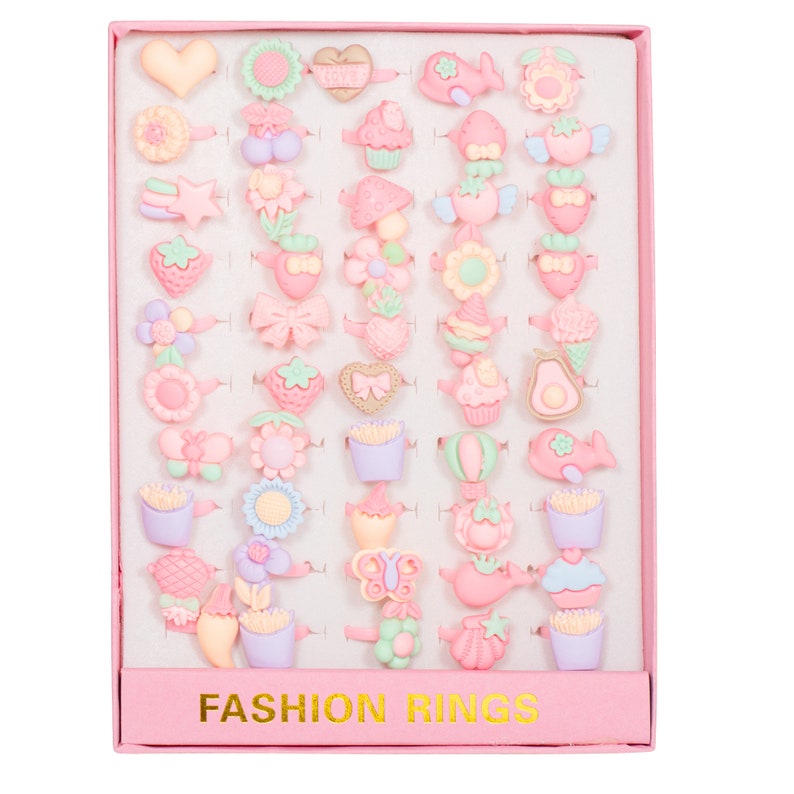 May include: A pink box with a white interior containing 36 colorful plastic rings with various designs, including hearts, flowers, cupcakes, and other cute motifs. The box has the words "FASHION RINGS" printed on the front.