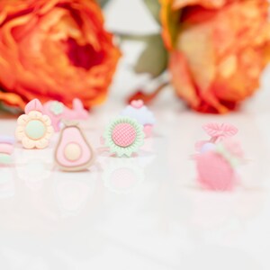 May include: A collection of colorful, small, plastic charms in the shape of a sunflower, avocado, cupcake, and a flower. The charms are on a white surface.
