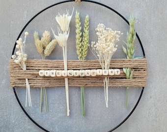 May include: A black metal hoop with a variety of dried flowers and grasses, including white, beige, and green. The hoop is decorated with a jute ribbon and wooden letter tiles spelling "HEREINS PAZIERET".
