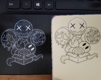 May include: A black and white sticker featuring a cartoon design. The design includes several circular shapes with different faces and patterns, and a box with more cartoon faces. The sticker is on a black surface.
