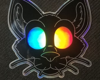 May include: A black sticker featuring a stylized cat face with large, colorful eyes. The eyes have a rainbow gradient, transitioning from blue to yellow and orange. The sticker has a holographic, reflective outline.