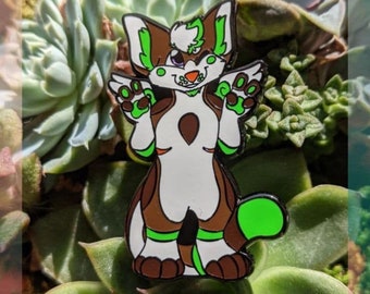 May include: A colorful enamel pin featuring a cartoon fox-like character with brown, white, and green accents. The pin has the text "ATLAS #014" and "THAT ONE PINK DOG STUDIOS". The character has its arms up in a welcoming gesture.