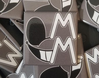 May include: Square gray buttons with a cartoon face design. The design features a smiling face with black eyes and a white "M" shape. The buttons are scattered, showing the front and sides of the buttons.