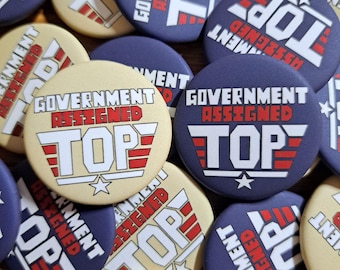 May include: A collection of round buttons with the text "GOVERNMENT ASSIGNED TOP" in white and red on a navy blue and cream background. Each button features a star and a stylized wing design.