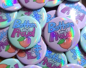 May include: A collection of round, holographic buttons with the text "Bottom and Proud" in blue and pink, and a peach illustration. The buttons have a light pink and mint green background.