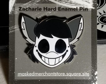 May include: Zacharie hard enamel pin featuring a stylized character with a large grin, black hair, and gray ears. The pin is on a gray card with the text "Zacharie Hard Enamel Pin" and "maskedmerchantstore.square.site".