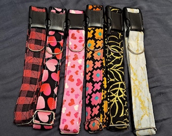 May include: A collection of six dog collars with various patterns. The collars feature black plastic buckles and metal D-rings. Patterns include red plaid, hearts on black, pink hearts, geometric shapes, gold leaf on black, and gold glitter on white.