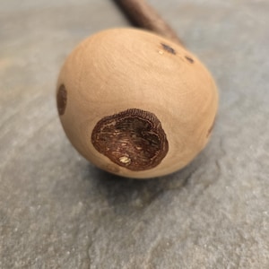 May include: A light-coloured wooden object with a rounded head and a long, thin handle. The head features dark brown, textured spots. The object is resting on a grey, textured surface.