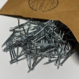 Puede incluir: Primer plano de un montón de clavos de color plateado. Los clavos están dispuestos en un patrón aleatorio y todos son del mismo tamaño. Los clavos están en una bolsa de papel marrón con el texto "OMO STUDIO" impreso en ella.