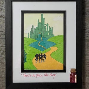 Può includere: Un'illustrazione incorniciata della Città di Smeraldo del Mago di Oz. I personaggi camminano lungo la strada di mattoni gialli verso la città. La citazione "Non c'è posto come casa" è scritta sotto l'immagine. Una piccola bottiglia di glitter è attaccata alla cornice.