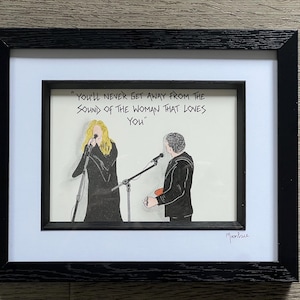 May include: A black and white drawing of two people singing on stage. The drawing is framed in a black frame with a white mat. The text "YOU'LL NEVER GET AWAY FROM THE SOUND OF THE WOMAN THAT LOVES YOU" is written in black ink above the drawing. The artist's signature "Moonface" is written in black ink in the bottom right corner of the drawing.
