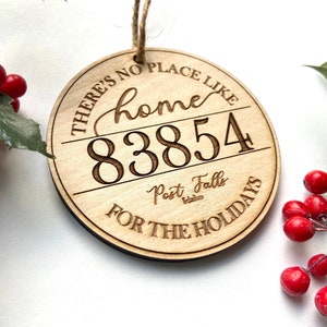 Könnte beinhalten: Ein runder Holzanhänger mit dem Text "There's no place like home 83854 Post Falls Idaho for the holidays".