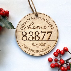 Puede incluir: Un adorno navideño de madera con el texto "There's no place like home 83877 Post Falls Idaho for the holidays".