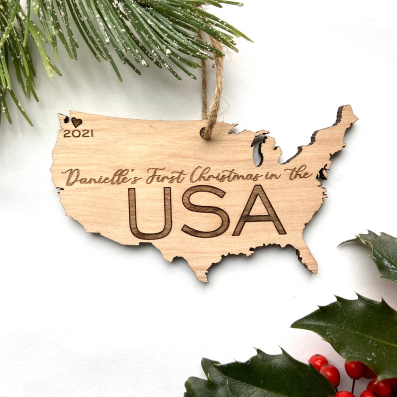 May include: A light brown USA-shaped wooden Christmas ornament.  The ornament features the text 'Danielle's First Christmas in the USA' and '2021' with a small heart symbol. The text is laser-engraved.  The ornament has a jute twine hanger. This keepsake is perfect for celebrating a first Christmas in the United States.
