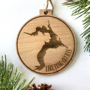 May include: A round wooden ornament with a laser-cut design of Lake Pend Oreille, a lake in Idaho. The ornament is light brown and has a rustic look.
