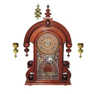 Puede incluir: Un reloj antiguo de madera con esfera redonda y detalles ornamentados. El reloj tiene un acabado marrón oscuro con detalles dorados y elementos decorativos. Dos candelabros dorados a cada lado.