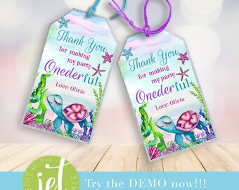 Può includere: Due tag di ringraziamento turchese e viola con sfondo glitter argentato. I tag raffigurano una tartaruga marina di cartone animato con cuori rosa sul guscio, circondata da alghe marine, conchiglie e stelle marine. Il testo sui tag recita "Grazie per aver reso la mia festa Onederful" con il nome Olivia.