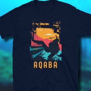 May include: A navy blue t-shirt featuring a retro-style graphic of a scuba diver in the water. The graphic depicts a sunset with orange, yellow, and pink colours. The text "AQABA" is printed in yellow below the graphic.