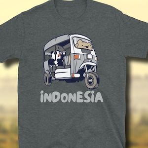 Op de afbeelding: Een grijs T-shirt met een cartoonillustratie van een wit en grijs driewielig voertuig met twee panda's erin. De tekst "INDONESIA" is onder het voertuig afgedrukt.