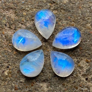 May include: Five teardrop-shaped moonstone cabochons with iridescent blue and white colors arranged in a flower pattern on a gray textured surface.
