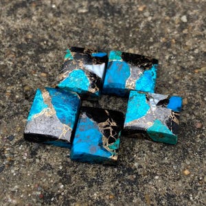 Könnte beinhalten: Sechs quadratische türkisfarbene Cabochons mit schwarzer und goldener Marmorierung. Die Cabochons sind in einem Halbkreis auf einer grauen, strukturierten Oberfläche angeordnet.