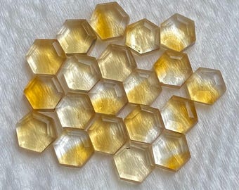 May include: A collection of faceted, hexagonal gemstones in shades of yellow and gold. The gems are arranged on a white, textured surface, with light reflecting off their cut surfaces. The overall effect is one of warmth and subtle sparkle.