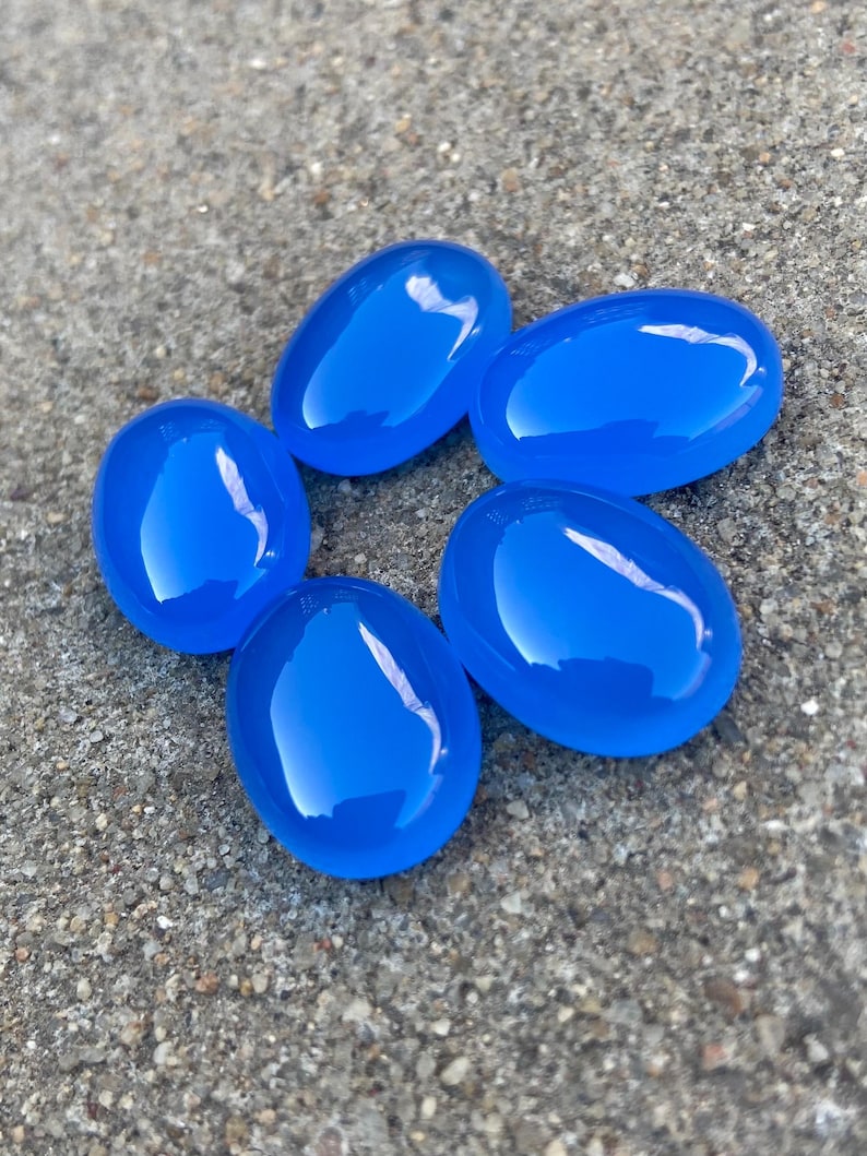 May include: Six smooth, oval-shaped blue gemstones. The stones are arranged in a circle on a gray, textured surface.