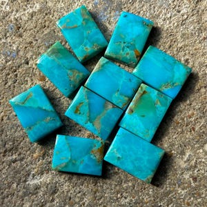 Peut inclure: Dix cabochons en turquoise, chacun de forme rectangulaire et de couleur bleu-vert avec des veines brunes. Les pierres sont disposées en un tas lâche sur une surface en béton gris.