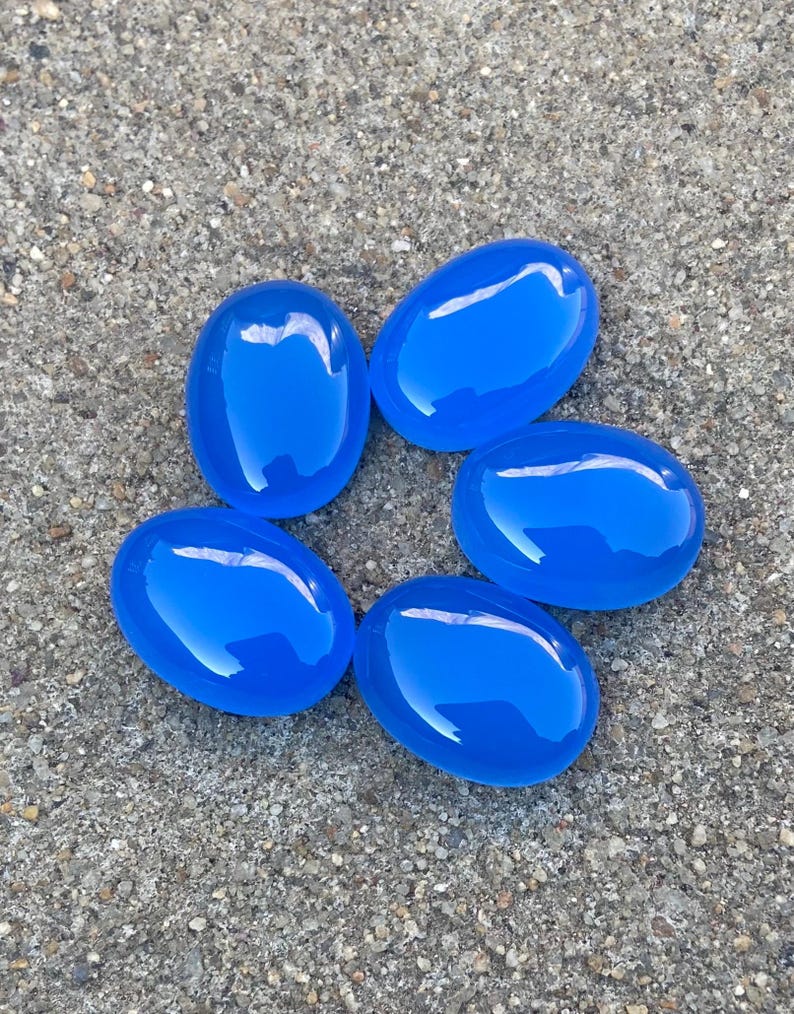 May include: Six smooth, oval-shaped, bright blue gemstones. The stones are arranged in a circle on a gray, textured surface.