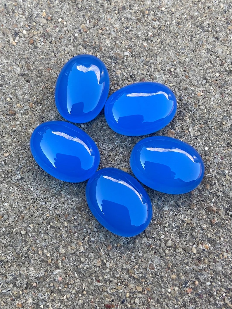 May include: Five smooth, oval-shaped blue gemstones. The stones are arranged in a circle on a gray, textured surface.