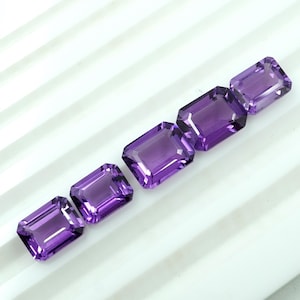 Puede incluir: Cinco piedras preciosas de amatista de corte rectangular en varios tonos de púrpura están dispuestas en diagonal. Las gemas tienen una superficie pulida y están sobre un fondo blanco y acanalado. Las piedras son de tamaño y forma similares.