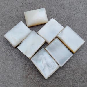 Könnte beinhalten: Sieben rechteckige, schillernde Perlmuttfliesen auf einer grauen Oberfläche. Die Fliesen haben eine perlmuttweiße Farbe mit subtilen Variationen und einem glänzenden Finish. Die Anordnung deutet auf ein dekoratives Element hin.