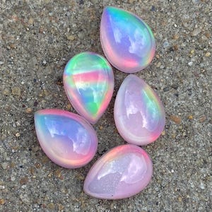 Puede incluir: Cinco piedras de cristal iridiscentes en forma de lágrima en tonos de rosa, morado y azul. Las piedras tienen un acabado nacarado y están dispuestas sobre una superficie gris texturizada.