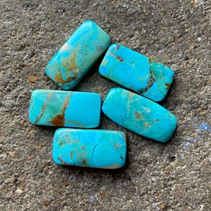 Könnte beinhalten: Fünf türkisfarbene Cabochons, jeder mit einer rechteckigen Form und einem einzigartigen Muster aus braunen und blauen Farbtönen. Die Steine sind auf einer rauen, grauen Oberfläche angeordnet.