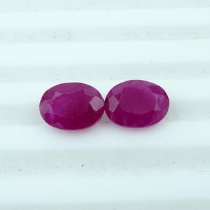 以下が含まれることがあります： 深紅色のオーバルカットルビー2つ。白い表面に展示されています。ルビーはファセットカットで、光を反射し、豊かな色合いを際立たせています。ジュエリー作りやコレクションに最適です。