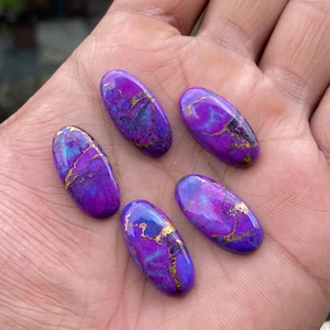 May include: Five oval-shaped purple gemstones with gold veining. The stones have a polished surface and a mix of purple, blue, and gold colors. The gemstones are displayed on a light-colored background.