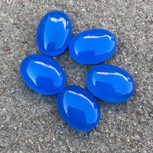 May include: Five smooth, oval-shaped blue gemstones. The stones are arranged in a circle on a gray, textured surface.