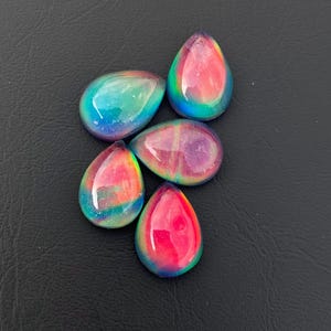 May include: Five iridescent teardrop-shaped cabochons with a rainbow of colors, including pink, blue, green, and purple. The cabochons are arranged in a cluster on a black surface.