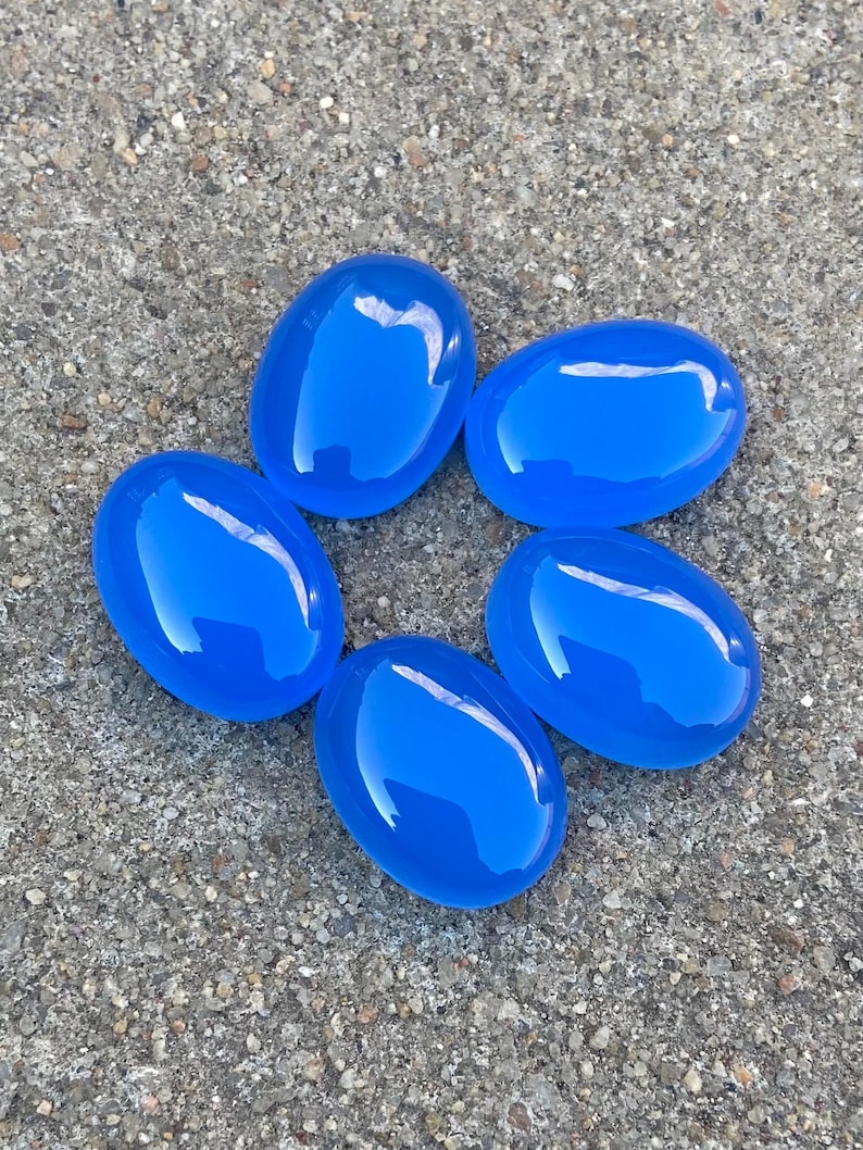 May include: Five smooth, oval-shaped blue gemstones arranged in a circle on a gray, textured surface.