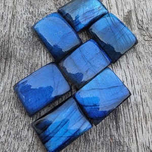 May include: Seven polished, dark blue labradorite stones, each cut into a rectangular shape, arranged in a staggered pattern on a wooden surface.