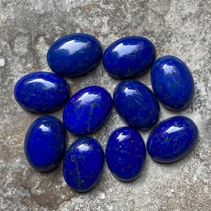 May include: Ten oval-shaped lapis lazuli stones, each with a deep blue colour and a slightly different pattern of white veins. The stones are arranged in a loose cluster on a grey, textured surface.
