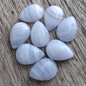 Pode incluir: Oito pedras preciosas polidas de ágata rendada azul em forma de gota. Cada pedra tem um tom azul pálido com padrões em redemoinho e listrados. As pedras são exibidas em uma superfície de madeira envelhecida.