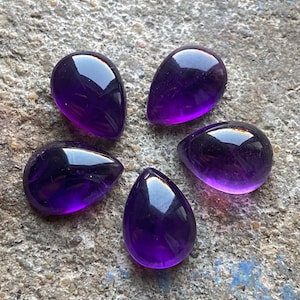 Può includere: Cinque pietre preziose di ametista a forma di goccia, ciascuna con una tonalità viola profonda e una finitura lucida. Le pietre sono disposte a forma di stella su una superficie grigia testurizzata.