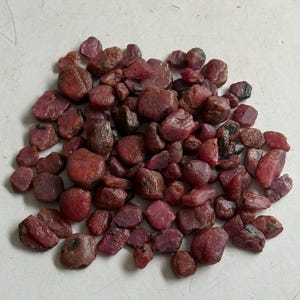 Könnte beinhalten: Eine Ansammlung zahlreicher roher, unpolierter Rubin-Edelsteine in verschiedenen Schattierungen von tiefem Rot und Rosa. Diese Steine weisen unregelmäßige, natürliche Formen mit texturierten, matten Oberflächen und einigen sichtbaren dunklen Einschlüssen auf.