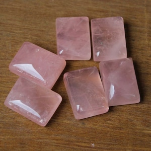 May include: Six rectangular rose quartz stones, each with a smooth, polished surface and a pale pink colour. The stones are arranged on a wooden surface.